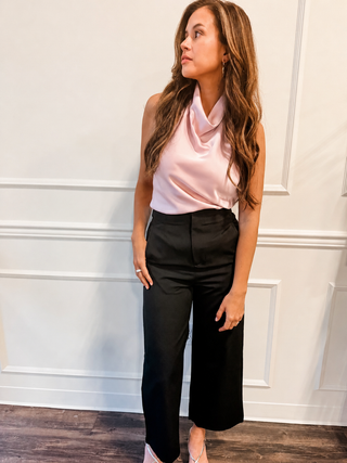 Woman wearing a pink sleeveless top and black pants against a white paneled wall.