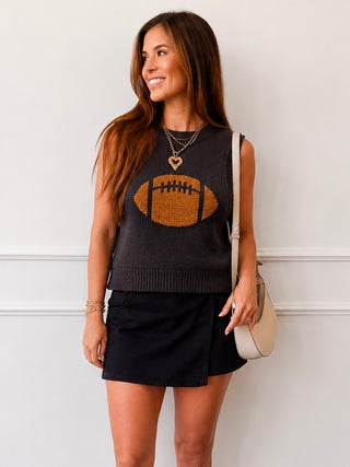 A woman wearing a dark charcoal sleeveless knit sweater vest with a brown football graphic on the front, paired with a black mini skirt. She is holding a beige shoulder bag and wearing gold necklaces, standing against a white wall with molding.