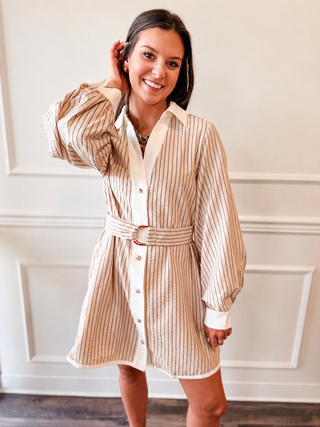 A woman modeling a beige and white vertically striped shirt dress with a matching belt and white collar, set against a white paneled wall.