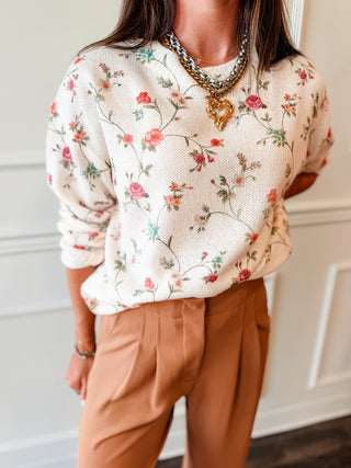 A woman wearing a cream-colored knit sweater with a delicate pink, red, and green floral pattern, paired with brown trousers and layered gold necklaces.