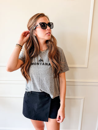 A woman wearing a heather gray graphic t-shirt with 'MONTANA' printed on the chest, paired with a black denim mini skirt and sunglasses, posing against a white paneled wall.