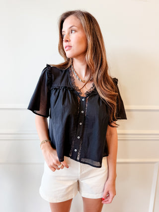 A woman wearing a black short-sleeve blouse with sheer ruffle sleeves and lace trim, paired with white shorts, standing against a white paneled wall.