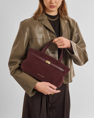 A model wearing a metallic olive-brown leather jacket and black top holds a burgundy suede handbag with a gold-tone turn-lock closure and gold logo text. The background is a clean, neutral gray studio backdrop.