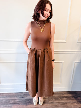 A woman wearing a sleeveless brown mock-neck top and matching pleated midi skirt with side pockets, standing in front of a white paneled wall.