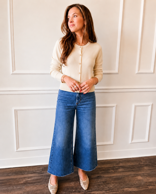 A woman modeling a cream-colored long-sleeve button-up cardigan and wide-leg blue denim jeans, standing in front of a white paneled wall on a dark wood floor.