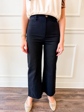 A person wearing black wide-leg cropped pants with a cream sleeveless top and beige woven pointed flats, standing against a white paneled wall.