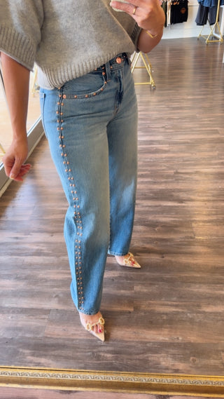 A person wearing light blue denim jeans with decorative gold-tone studs down the front seams, paired with a gray knit sweater and pointed shoes, standing in a boutique with a wood floor.