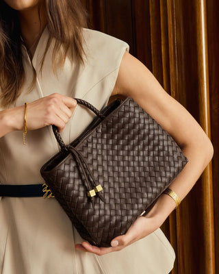 A woman in a beige sleeveless dress holds a dark brown woven leather handbag with gold-tone hardware against a warm wood-paneled background.