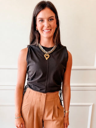 A woman wearing a sleeveless black satin top with a gold heart pendant necklace and tan high-waisted trousers, standing against a white paneled wall.