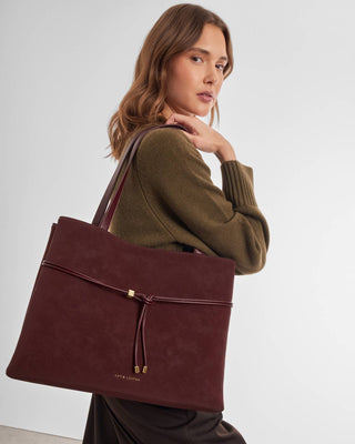 A woman wearing an olive green sweater carries a large burgundy suede tote bag with a drawstring closure and gold-tone hardware against a plain light gray studio background.