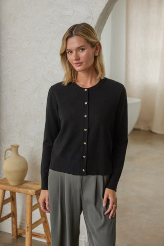 A woman wearing a black long-sleeve button-up cardigan with gold-tone buttons and gray trousers, standing in a softly lit indoor setting with a textured wall, stool, and ceramic jug.