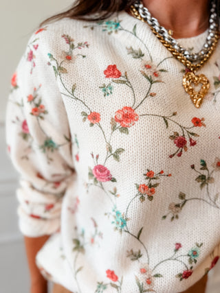 A close-up of a person wearing a cream knit sweater with a delicate floral vine pattern in pink, red, and green. The person is also wearing layered gold and silver chain necklaces with a gold heart pendant.