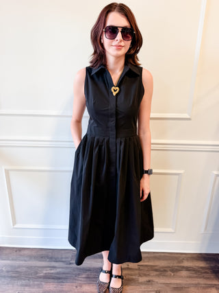 A woman wearing a sleeveless black midi dress with a button-down collar, a gathered waist, and a flared skirt, styled with sunglasses, a gold heart-shaped pendant, and black-and-white patterned heels, standing against a white paneled wall.
