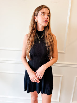 A woman with long light brown hair wearing a sleeveless black ribbed mini dress with a scalloped hem, standing against a white paneled wall.