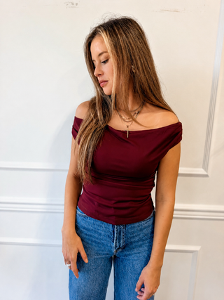 A woman wearing a fitted off-the-shoulder burgundy top and blue jeans, standing against a white paneled wall.