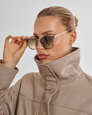 A close-up of a woman wearing stylish cat-eye sunglasses with gold frames and gradient lenses, paired with a high-collar taupe faux-leather jacket against a light gray background.