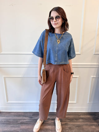 A woman modeling a casual outfit consisting of a blue cropped short-sleeve denim top, brown wide-leg pants, white sneakers, and a brown crossbody bag, standing against a white paneled wall.
