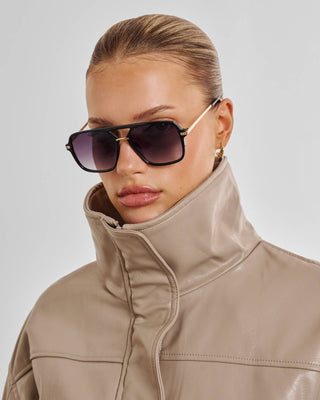 Woman wearing a beige high-neck jacket and dark sunglasses against a light gray background