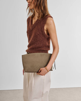 A woman wearing a brown fuzzy sleeveless sweater and cream trousers holds a large taupe suede clutch bag against a plain light gray studio background.