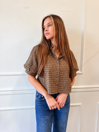 A woman with long brown hair wearing a brown and beige plaid short-sleeve button-up shirt and blue jeans, standing against a white paneled wall.