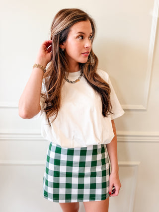 A woman wearing a cream-colored short-sleeve top and a green and white gingham mini skirt, standing against a white paneled wall.