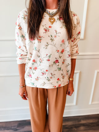 A woman wearing a cream-colored waffle-knit sweater with a red and green floral pattern, paired with tan pants, standing in front of a white paneled wall.
