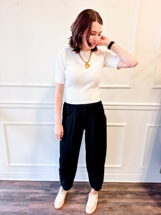 A woman modeling a white short-sleeve knit top and black wide-leg pants, paired with white sneakers and a gold necklace, standing against a white paneled wall.