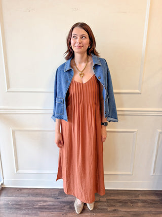 A woman stands indoors wearing a rust-orange sleeveless maxi dress with a pleated texture, layered under a light blue denim jacket. She is standing in front of a white paneled wall on a dark wood floor.