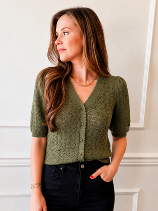 A woman wearing an olive green short-sleeve knit cardigan with a scalloped V-neck and button-front closure, paired with black jeans, standing against a white paneled wall.