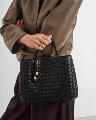 A person wearing a brown leather jacket holds a black woven leather handbag with gold-tone hardware against a plain light background.