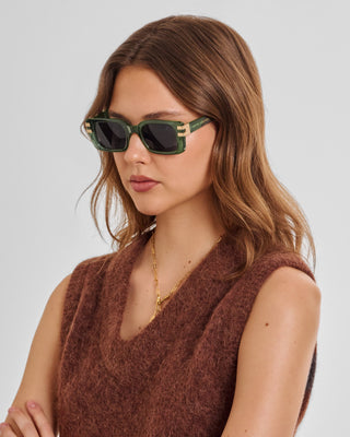 A woman with wavy brown hair wearing green square sunglasses with gold geometric accents and a brown knit sleeveless top, photographed against a light gray studio background.